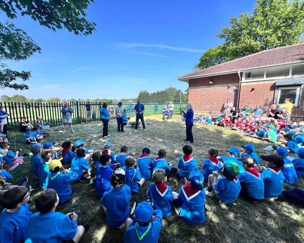 Beavers and Cubs from across the district sitting on the grass outside a Scout hut, listening to their leaders.