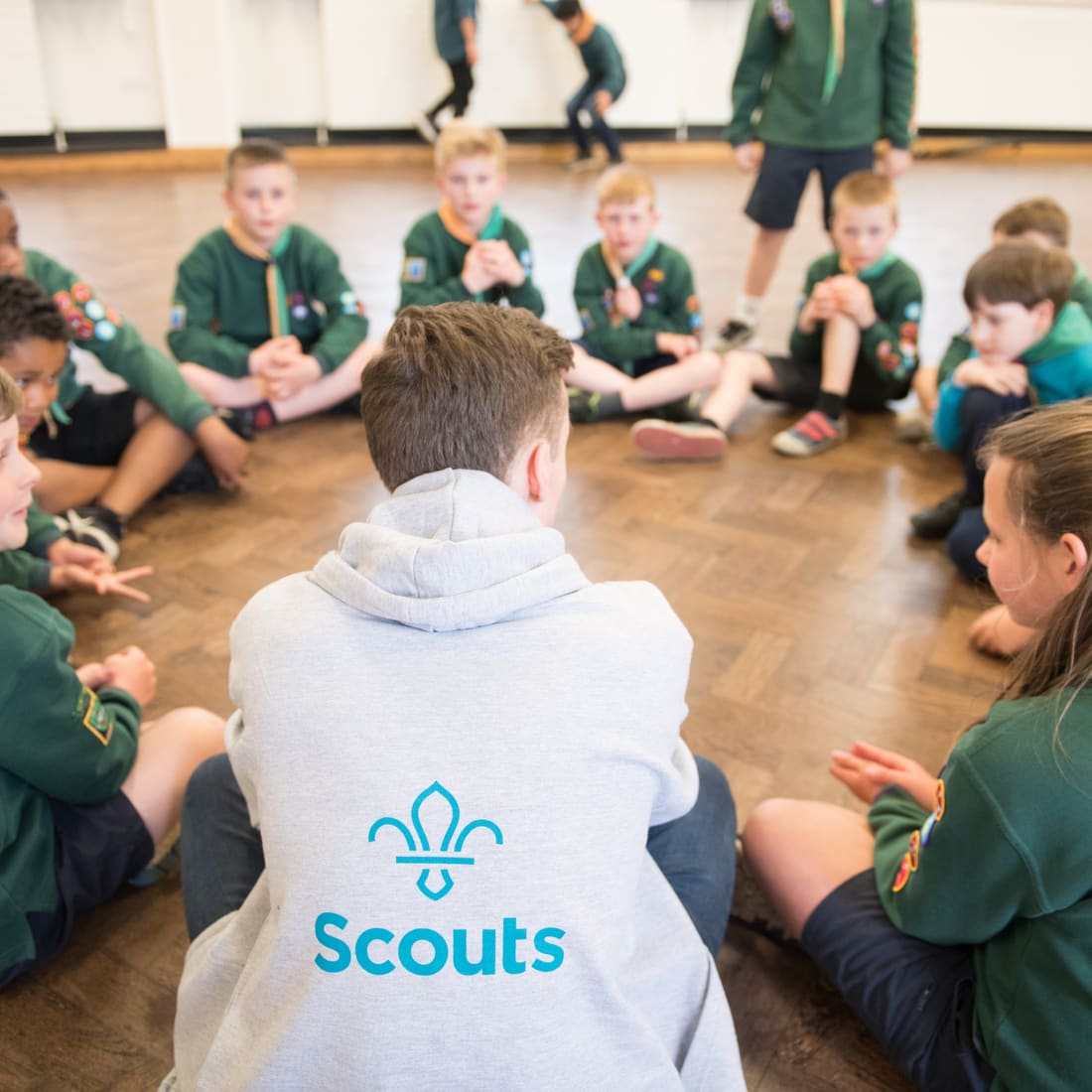 Cubs in green jumpers sitting cross-legged in a circle on a hall floor, listening to a volunteer in a Scouts hoodie sitting among them