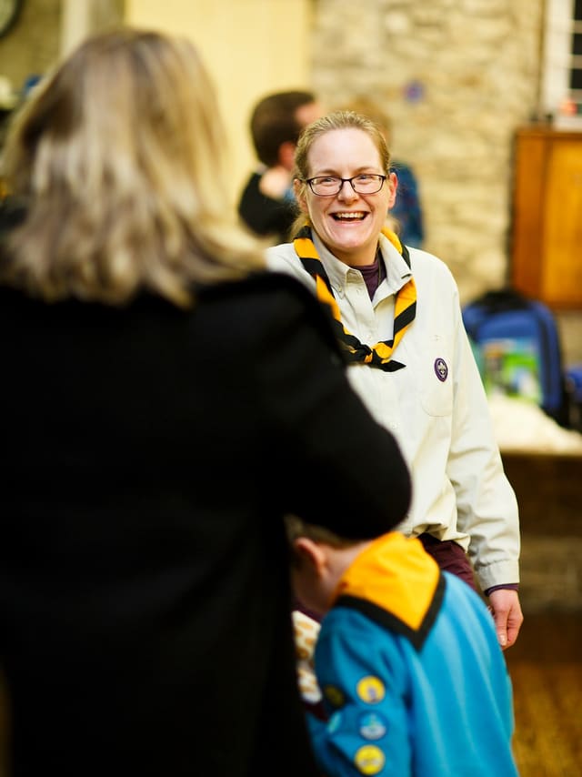 A Scout leader in uniform laughing while greeting a parent at the start of a section night, with a young person in a Beavers jumper arriving in front of them