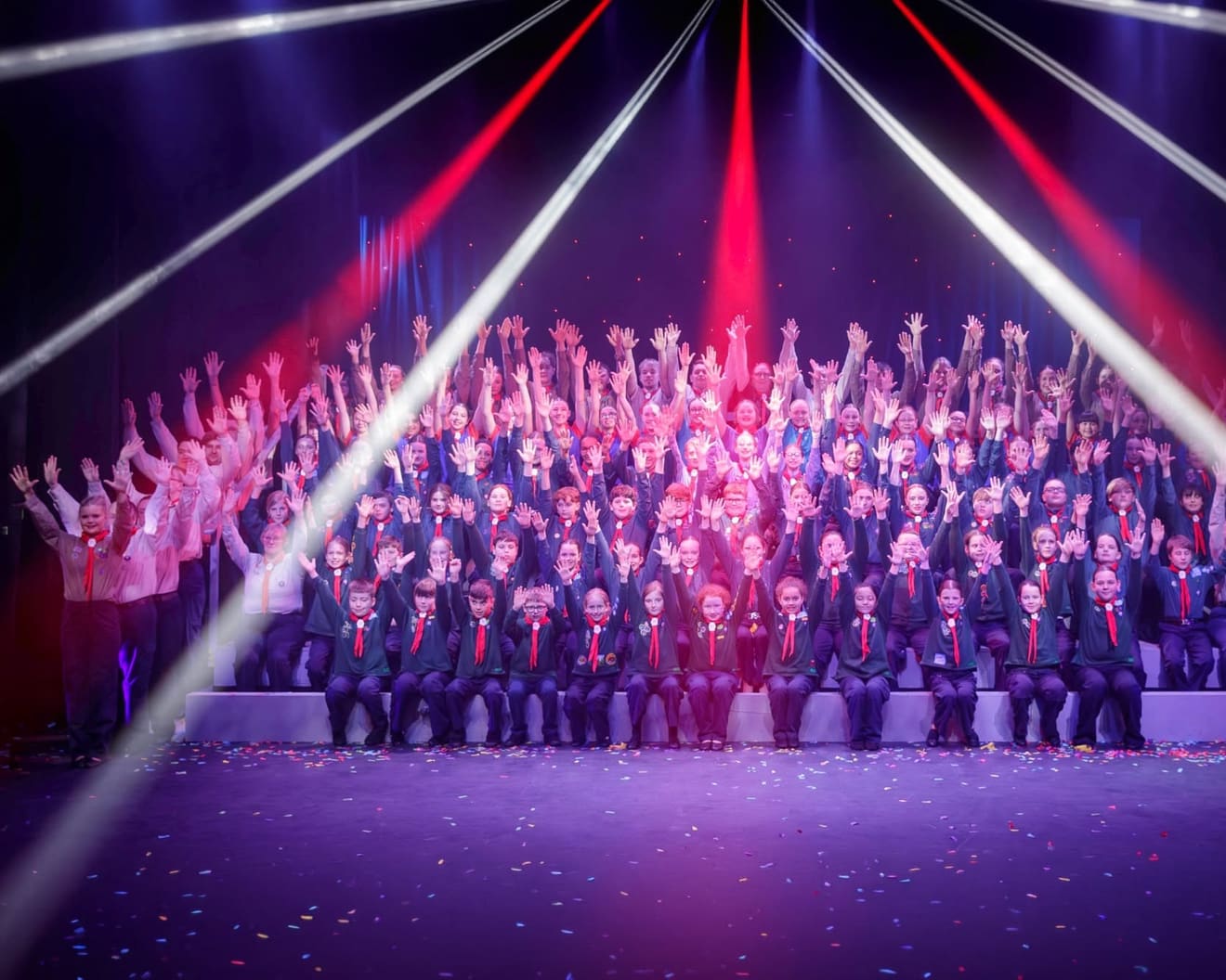 Scouts and volunteers from across the district on stage together, hands raised under stage lights and falling confetti.