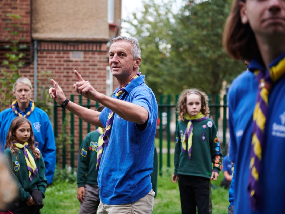 A volunteer in a Scouts polo shirt talking and gesturing, with Cubs and other volunteers listening